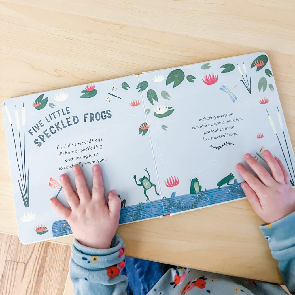Child's hands holding down open board book pages with text that reads "Five Little Speckled Frogs" and images of frogs hopping