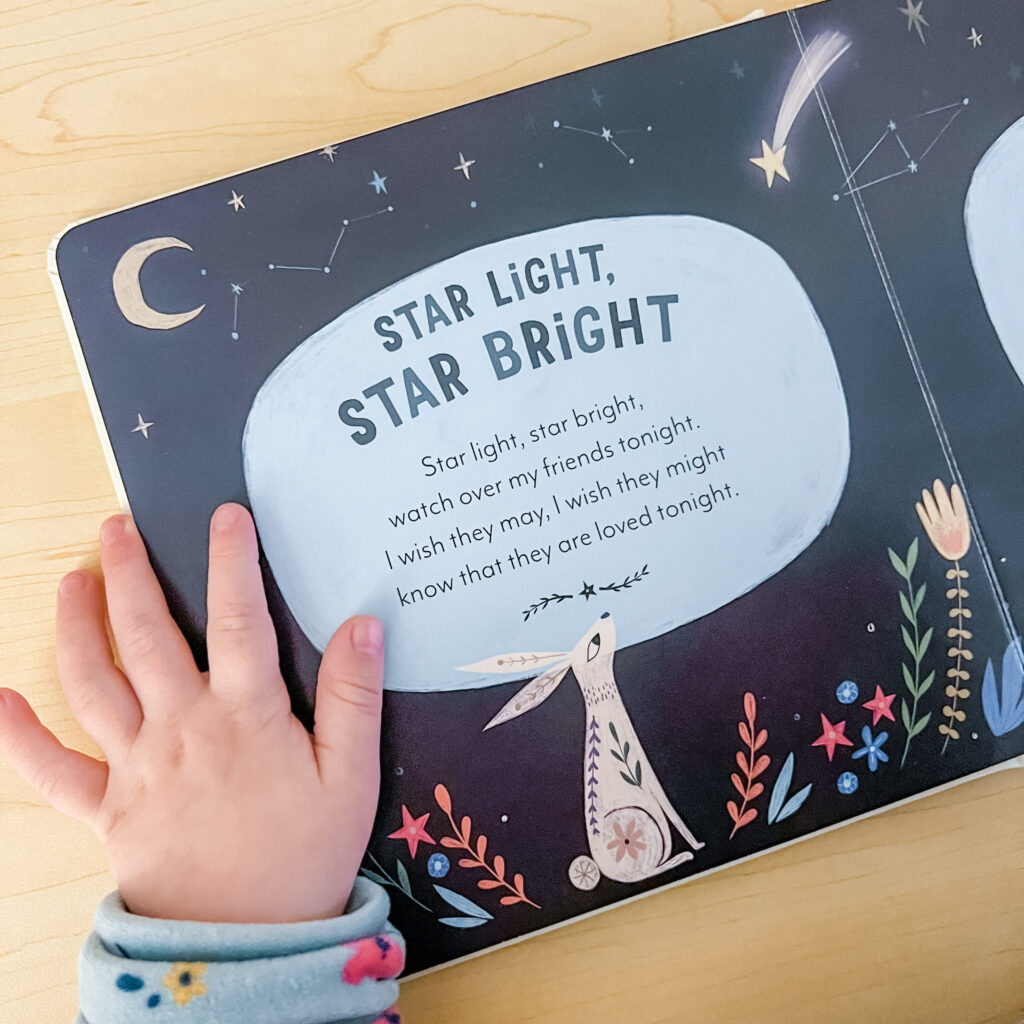 Child's hand holding down page of board book with night sky scene including moon and stars and pink rabbit looking up at text which reads Star Light, Star Bright