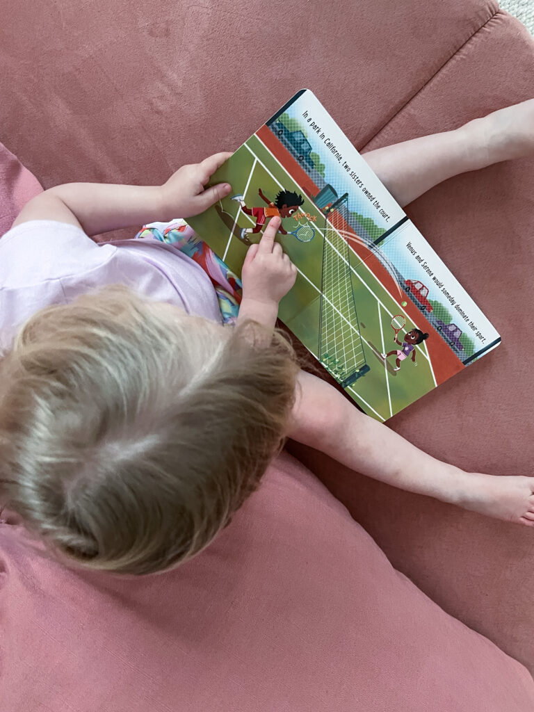 Child pointing to picture of girl playing tennis in board book Baby Ballers: Venus and Serena Williams